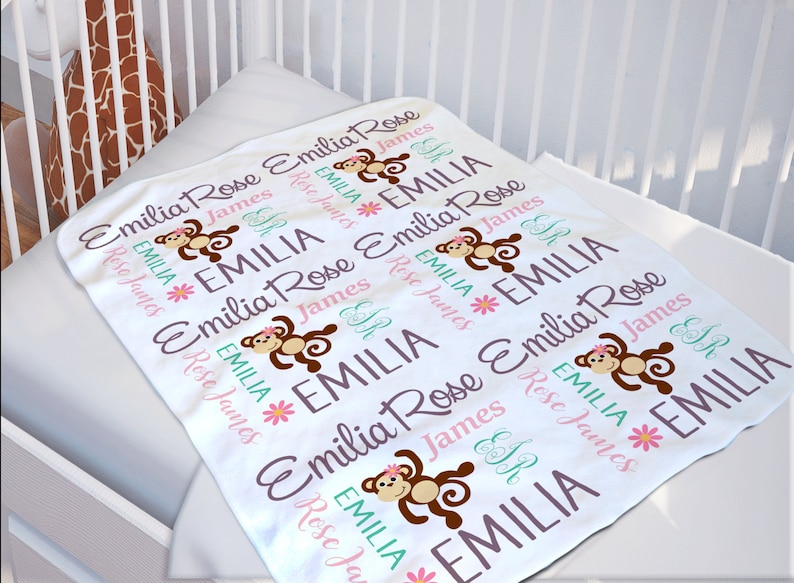 K&ouml;nnte beinhalten: Wei&szlig;e Babydecke mit einem sich wiederholenden Muster aus braunen Affen, rosa Blumen und den Namen "Emilia Rose" und "James" in lila und gr&uuml;nem Text.