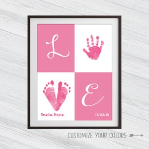 May include: Pink and white framed print with the word "LOVE" spelled out in white cursive letters. The "L" and "E" are on pink backgrounds, the "O" is a pink handprint on a white background, and the "V" is a pink footprint on a white background. The print also includes the name "Amelia Marie" and the date "12/02/16".