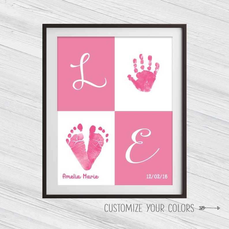 May include: Pink and white framed print with the word "LOVE" spelled out in white cursive letters. The "L" and "E" are on pink backgrounds, the "O" is a pink handprint on a white background, and the "V" is a pink footprint on a white background. The print also includes the name "Amelia Marie" and the date "12/02/16".