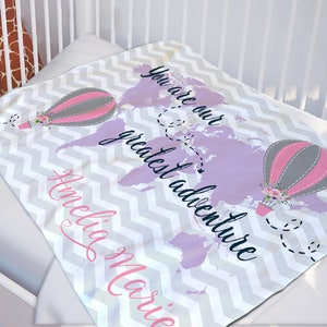 May include: A soft, gray and white chevron patterned baby blanket with a purple world map and two hot air balloons. The text "You are our greatest adventure" is written in black cursive font. The name "Amelia Marie" is written in pink cursive font.