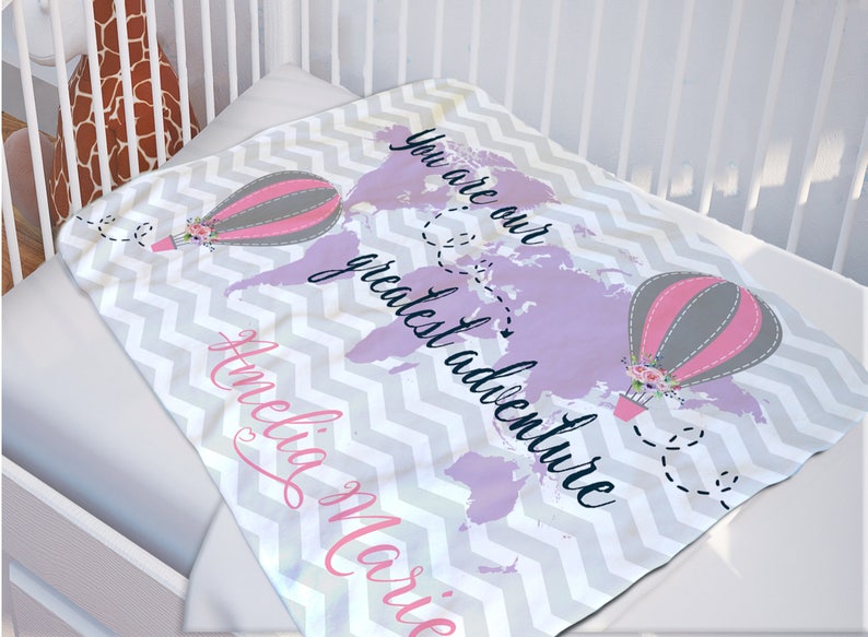 May include: A soft, gray and white chevron patterned baby blanket with a purple world map and two hot air balloons. The text "You are our greatest adventure" is written in black cursive font. The name "Amelia Marie" is written in pink cursive font.
