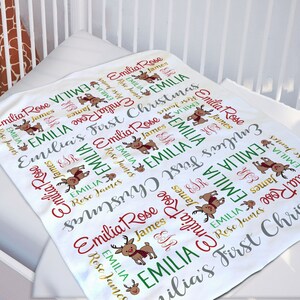May include: White baby blanket with a repeating pattern of reindeer and the text "Emilia's First Christmas". The reindeer are red, green, and brown. The text is in a variety of colors, including red, green, gold, and gray.