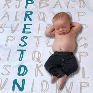 May include: A baby boy wearing blue jeans is lying on a white blanket with a teal alphabet pattern. The baby's name, "PRESTON", is spelled out vertically in teal letters.
