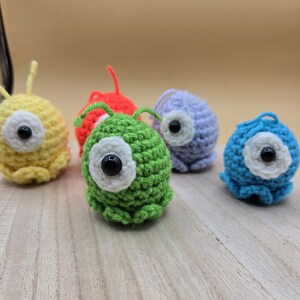 May include: A collection of six colorful, crocheted monster-like creatures. Each has a large, white-rimmed eye with a black center. The creatures are in shades of yellow, red, green, purple, and blue. They are arranged on a light-colored wooden surface.