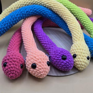 May include: A collection of colorful, crocheted snake toys in shades of pink, peach, purple, blue, yellow, and green. Each snake has black button eyes. The toys are arranged on a light brown surface.