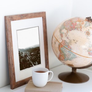 Pode incluir: Uma caneca de cer&acirc;mica branca com uma moldura de madeira marrom que cont&eacute;m uma fotografia em preto e branco de uma cordilheira. Tamb&eacute;m h&aacute; um globo terrestre e um caderno com um l&aacute;pis na prateleira branca.