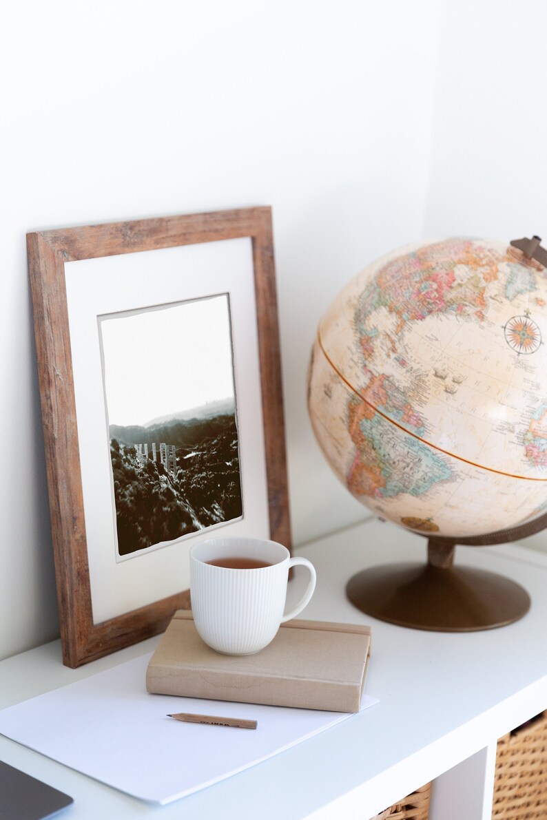 Pode incluir: Uma caneca de cer&acirc;mica branca com uma moldura de madeira marrom que cont&eacute;m uma fotografia em preto e branco de uma cordilheira. Tamb&eacute;m h&aacute; um globo terrestre e um caderno com um l&aacute;pis na prateleira branca.