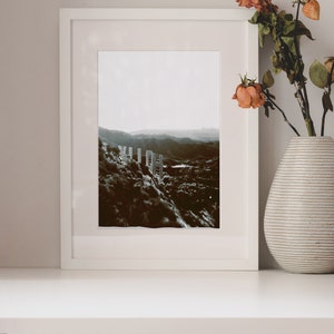 Pode incluir: Uma fotografia a preto e branco enquadrada em branco de uma paisagem com uma cordilheira de montanhas no fundo. A fotografia est&aacute; sobre uma prateleira branca com um vaso de flores secas e uma pilha de livros.
