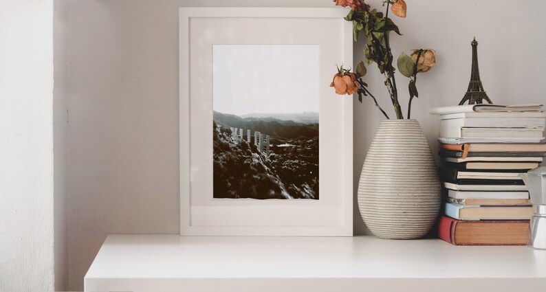Pode incluir: Uma fotografia a preto e branco enquadrada em branco de uma paisagem com uma cordilheira de montanhas no fundo. A fotografia est&aacute; sobre uma prateleira branca com um vaso de flores secas e uma pilha de livros.