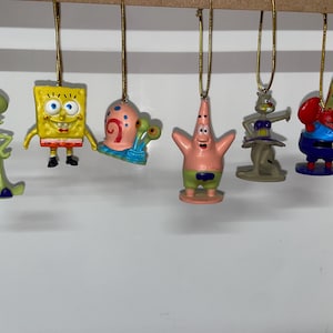 Op de afbeelding: Een verzameling van zes ornamenten van stripfiguren, waaronder SpongeBob SquarePants en Patrick Star. Elk ornament hangt aan een gouden draad. De karakters zijn helder gekleurd en gedetailleerd, perfect voor feestelijke decoraties.