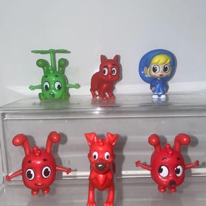 Puede incluir: Una colección de figuras de juguete coloridas. Los juguetes incluyen una figura verde con forma de alienígena con una hélice, un perro rojo, una niña con una capa con capucha azul y tres criaturas rojas con forma de insecto. Los juguetes están sobre una superficie transparente.