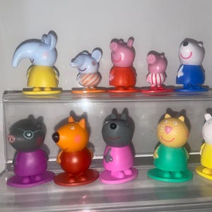 May include: A collection of colorful character figurines, including an elephant, mouse, pig, and bear. The figures are on a clear display shelf. Each figure has a unique design and color scheme, with details like clothing and facial expressions.