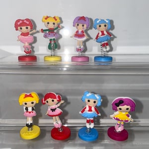 May include: Eight colorful, spring-loaded, plastic dolls with different outfits and hairstyles. The dolls are arranged in two rows of four.