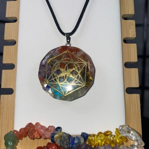 May include: A round, multi-coloured crystal pendant with a gold geometric design, hanging from a black cord. The pendant is surrounded by a variety of colourful tumbled stones.