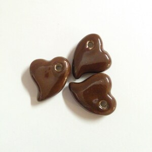 May include: Three glossy, brown heart-shaped ceramic beads. Each bead has a small hole at the top, suggesting they are designed for stringing or hanging. The beads are arranged on a white surface, with soft shadows.
