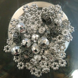 May include: A collection of silver and black metal beads, including round, faceted, and decorative beads. The beads are arranged in a cluster on a reflective surface.