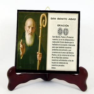 May include: A framed religious image depicting a bald man with a long white beard, holding a staff. The image is mounted on a dark wooden stand. Text in Spanish reads "San Benito Abad" and a prayer.