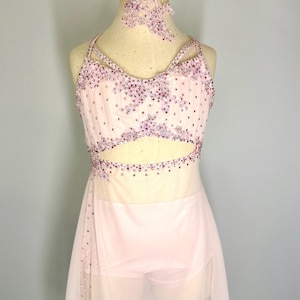 May include: A pale pink dance costume with a halter neck top and a skirt. The top and headband are decorated with purple rhinestones. The skirt is made of a sheer, flowing fabric. The costume is on a mannequin.