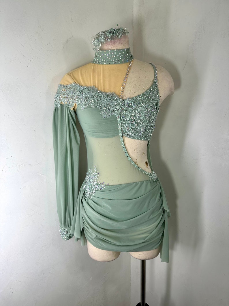 May include: A sage green dance costume with a sheer bodice, one long sleeve, and a gathered skirt. The bodice is embellished with lace and rhinestones.