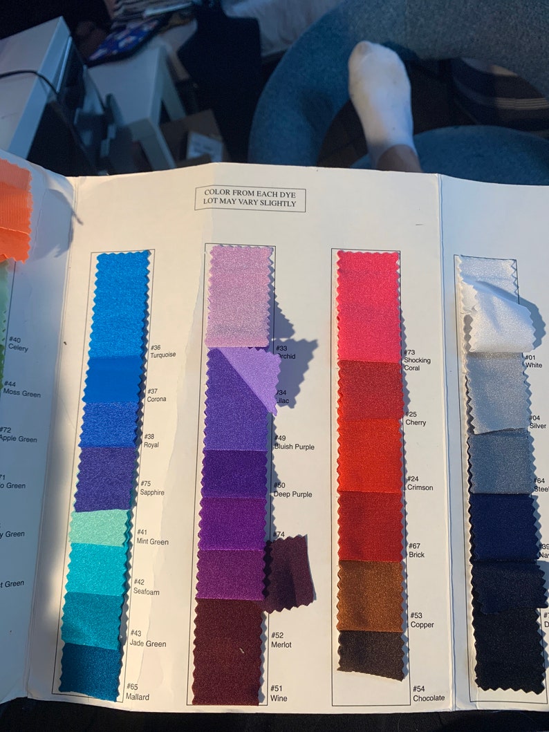 May include: A colour swatch book displaying a range of fabrics in shades of blue, purple, pink, red, and brown. The text "COLOR FROM EACH DYE LOT MAY VARY SLIGHTLY" is printed at the top. Each colour is labelled with a number and a name.