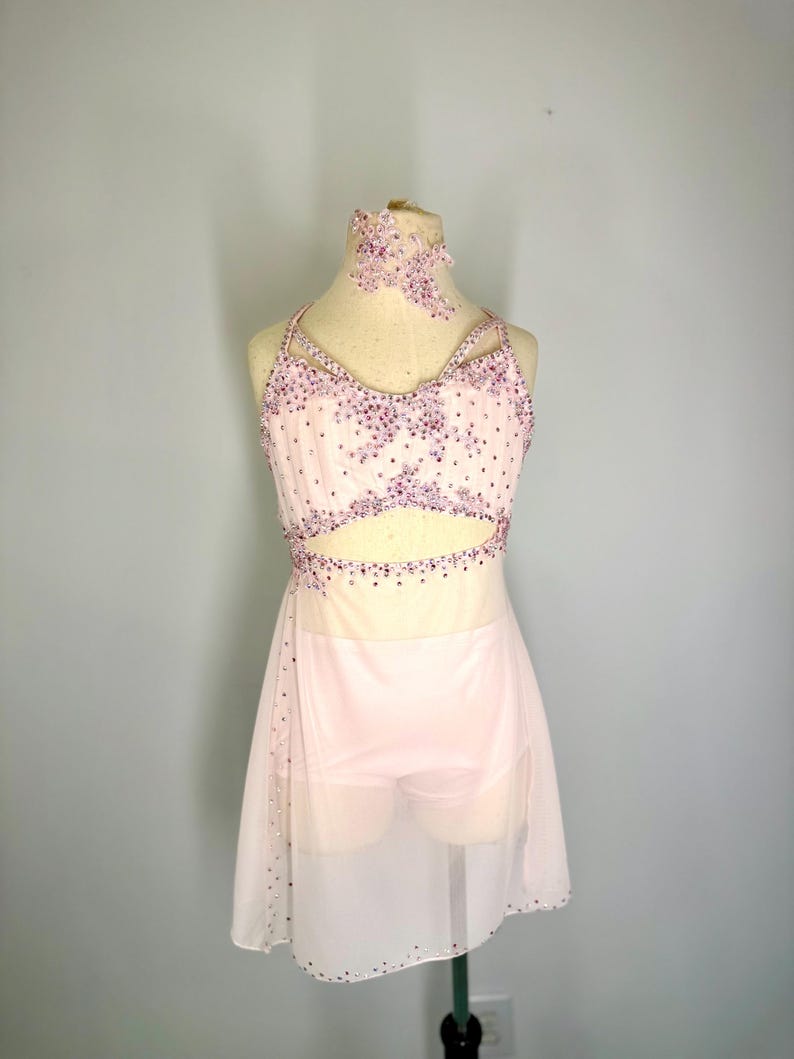 May include: A pale pink dance costume with a halter neck top and a skirt. The top and headband are decorated with purple rhinestones. The skirt is made of a sheer, flowing fabric. The costume is on a mannequin.