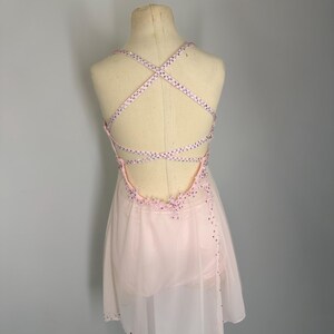 May include: A pale pink dance dress with a crisscross back. The dress has a sheer skirt and is decorated with sparkling embellishments on the neckline and straps. The dress is displayed on a mannequin.