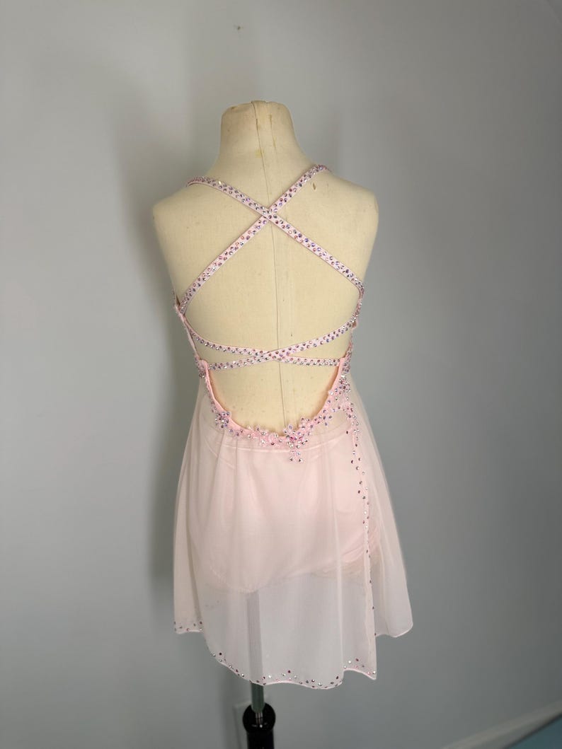 May include: A pale pink dance dress with a crisscross back. The dress has a sheer skirt and is decorated with sparkling embellishments on the neckline and straps. The dress is displayed on a mannequin.