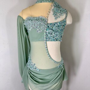May include: A sage green dance costume with a sheer bodice and long sleeves. The bodice is embellished with rhinestones and lace. The skirt is gathered and has a flowing silhouette.