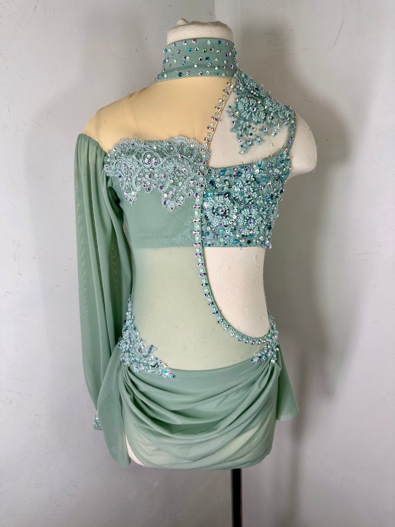 May include: A sage green dance costume with a sheer bodice and long sleeves. The bodice is embellished with rhinestones and lace. The skirt is gathered and has a flowing silhouette.