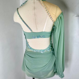 May include: A sage green dance costume with a one-shoulder design, featuring a long sheer sleeve with beaded embellishments. The costume has a fitted bodice with a low back and a flowing skirt.