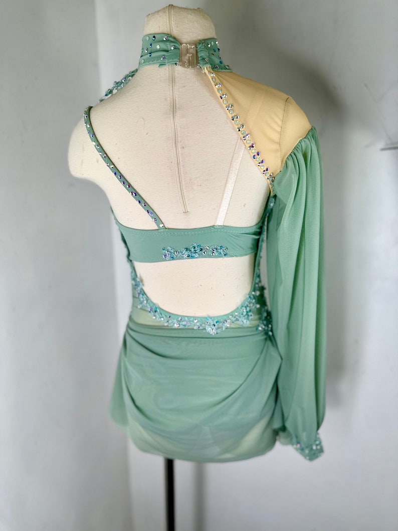 May include: A sage green dance costume with a one-shoulder design, featuring a long sheer sleeve with beaded embellishments. The costume has a fitted bodice with a low back and a flowing skirt.