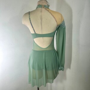 May include: A sage green dance costume with a sheer, long sleeve on one side and a sheer, sleeveless bodice with a cutout back. The costume is embellished with rhinestones and sequins.