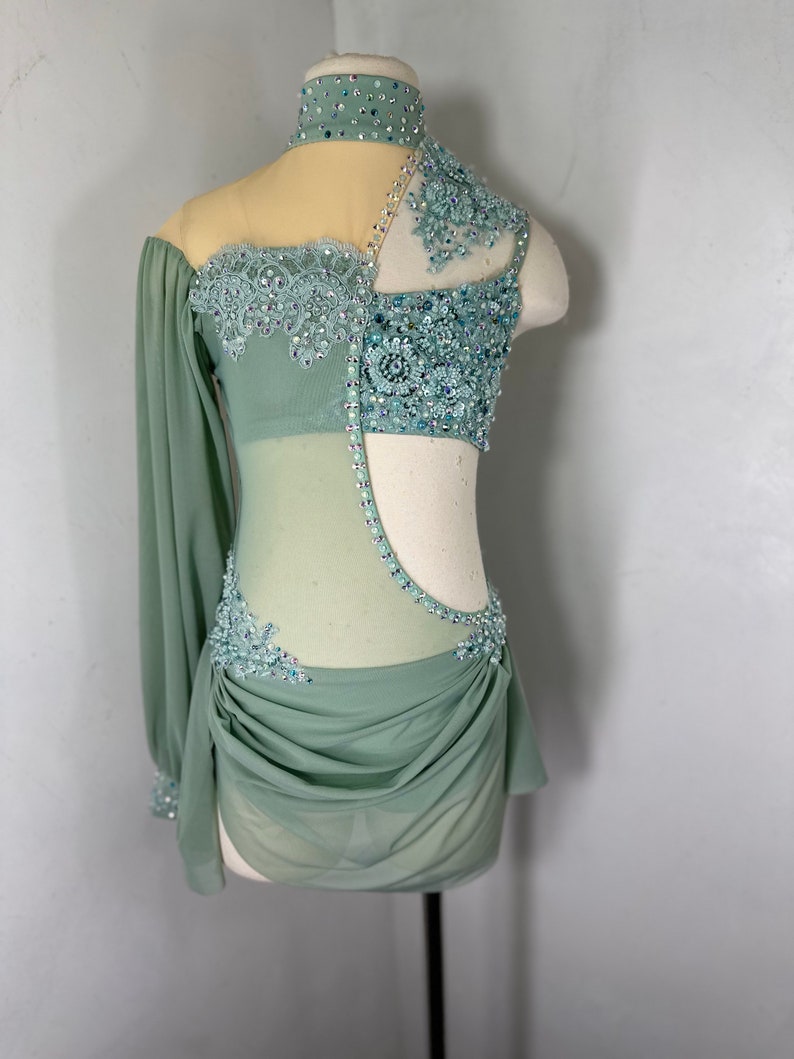 May include: A sage green dance costume with a sheer bodice and a long sleeve. The bodice is embellished with rhinestones and lace. The skirt is gathered and has a flowing drape.