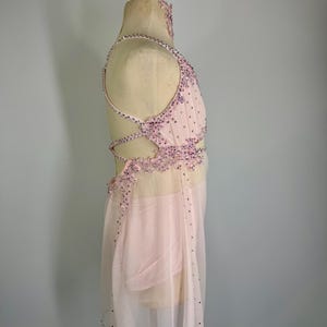 May include: A pale pink dance costume with detailed beadwork and a sheer skirt. The bodice has a halter neckline and crisscross back straps. Silver and purple rhinestones adorn the costume. It is displayed on a mannequin.