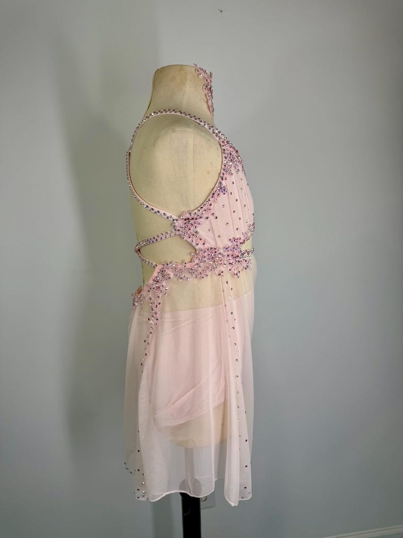 May include: A pale pink dance costume with detailed beadwork and a sheer skirt. The bodice has a halter neckline and crisscross back straps. Silver and purple rhinestones adorn the costume. It is displayed on a mannequin.