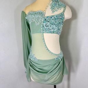 May include: A sage green dance costume with a sheer bodice and long sleeve. The bodice is embellished with rhinestones and lace. The skirt is gathered and flowing.