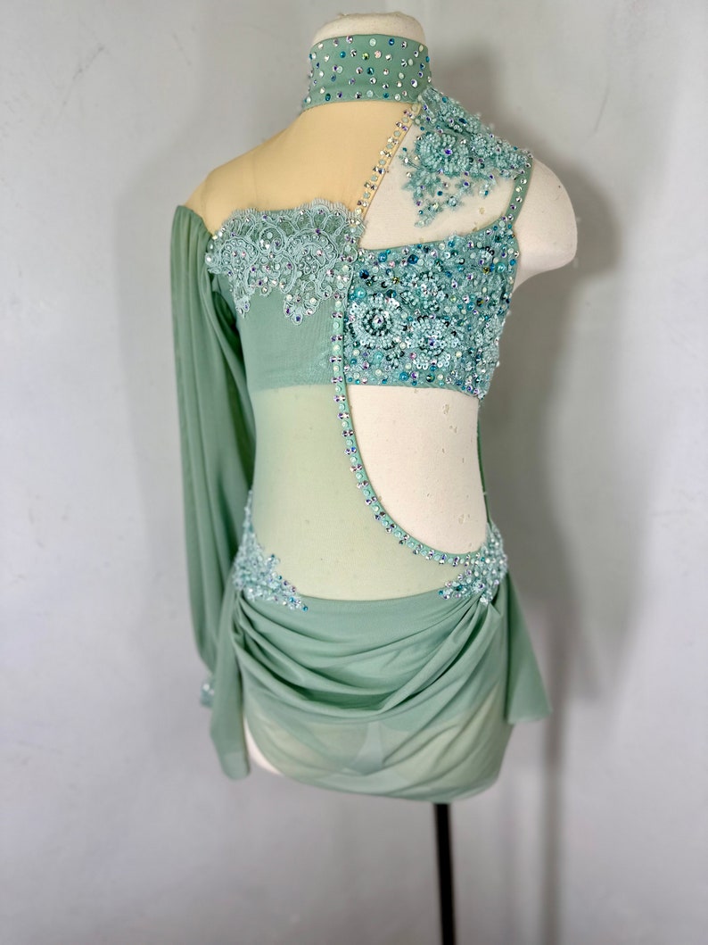 May include: A sage green dance costume with a sheer bodice and long sleeve. The bodice is embellished with rhinestones and lace. The skirt is gathered and flowing.