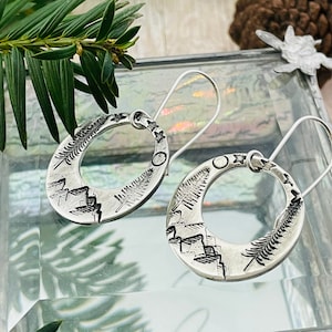 May include: A pair of silver oval hoop earrings. Each earring features a detailed landscape design with mountains, trees, and celestial elements. The earrings have a hook closure and are displayed on a glass surface.