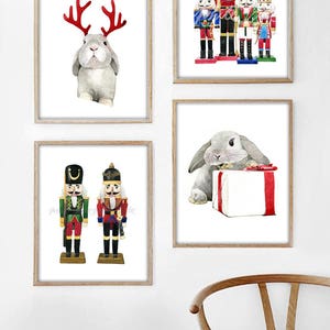 May include: Set of four watercolor prints featuring a gray rabbit wearing reindeer antlers, a gray rabbit holding a gift box, and two nutcracker soldiers. The prints are framed in a light wood frame.