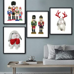 May include: A set of four framed prints featuring watercolor illustrations of nutcracker soldiers and bunnies wearing antlers and a red scarf. The prints are displayed on a wall above a couch with pillows.