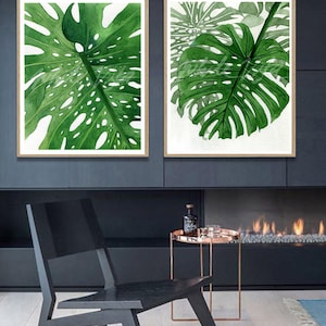 K&ouml;nnte beinhalten: Zwei gerahmte Aquarellbilder von gr&uuml;nen Monstera-Bl&auml;ttern. Die Bilder h&auml;ngen an einer dunkelgrauen Wand &uuml;ber einem Kamin.
