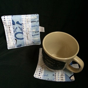 May include: Two blue and white fabric coasters with a black and white polka dot pattern. One coaster has a black chalkboard with the text "Seams Joyful" written on it. The other coaster has a blue and white fabric with text in black. A beige ceramic mug is sitting on the coaster with the chalkboard.