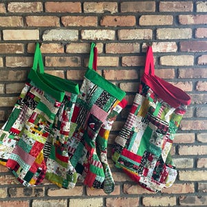 May include: Three Christmas stockings hang against a brick wall. The stockings are patchwork, with green, red, and white fabrics. Each stocking has a green or red cuff and a matching loop for hanging.