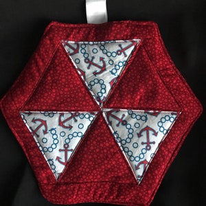 May include: Red and white quilted hot pad with a nautical theme. The pad is made of fabric with a red polka dot pattern and white fabric with blue anchors and chains. The pad is shaped like a hexagon.