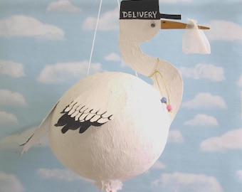 May include: A white stork ornament with a textured body, black wing details, and orange legs. The stork wears a black cap with the word DELIVERY printed in white and carries a small white fabric bundle in its beak, suspended by a white string.