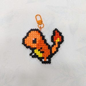May include: A cross-stitched Charmander keychain. The orange and yellow pixelated design features black outlines and a small orange clasp. The keychain is a handmade item.