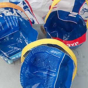May include: Three handmade blue and white tote bags with yellow handles. The bags are constructed from repurposed dog food packaging, featuring text and graphics. The bags are designed with a round shape.