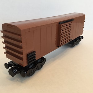 Puede incluir: Un vagón de mercancías de madera marrón con ruedas negras y una puerta de madera marrón con un diseño de listones. El vagón está hecho de ladrillos LEGO.