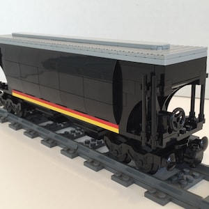 Puede incluir: Vagón de tren Lego negro con techo gris y rayas amarillas y rojas. El vagón está en una vía Lego gris.