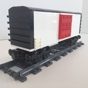 Puede incluir: Un vagón de tren Lego blanco y negro con una puerta roja. El vagón está en una vía Lego gris.